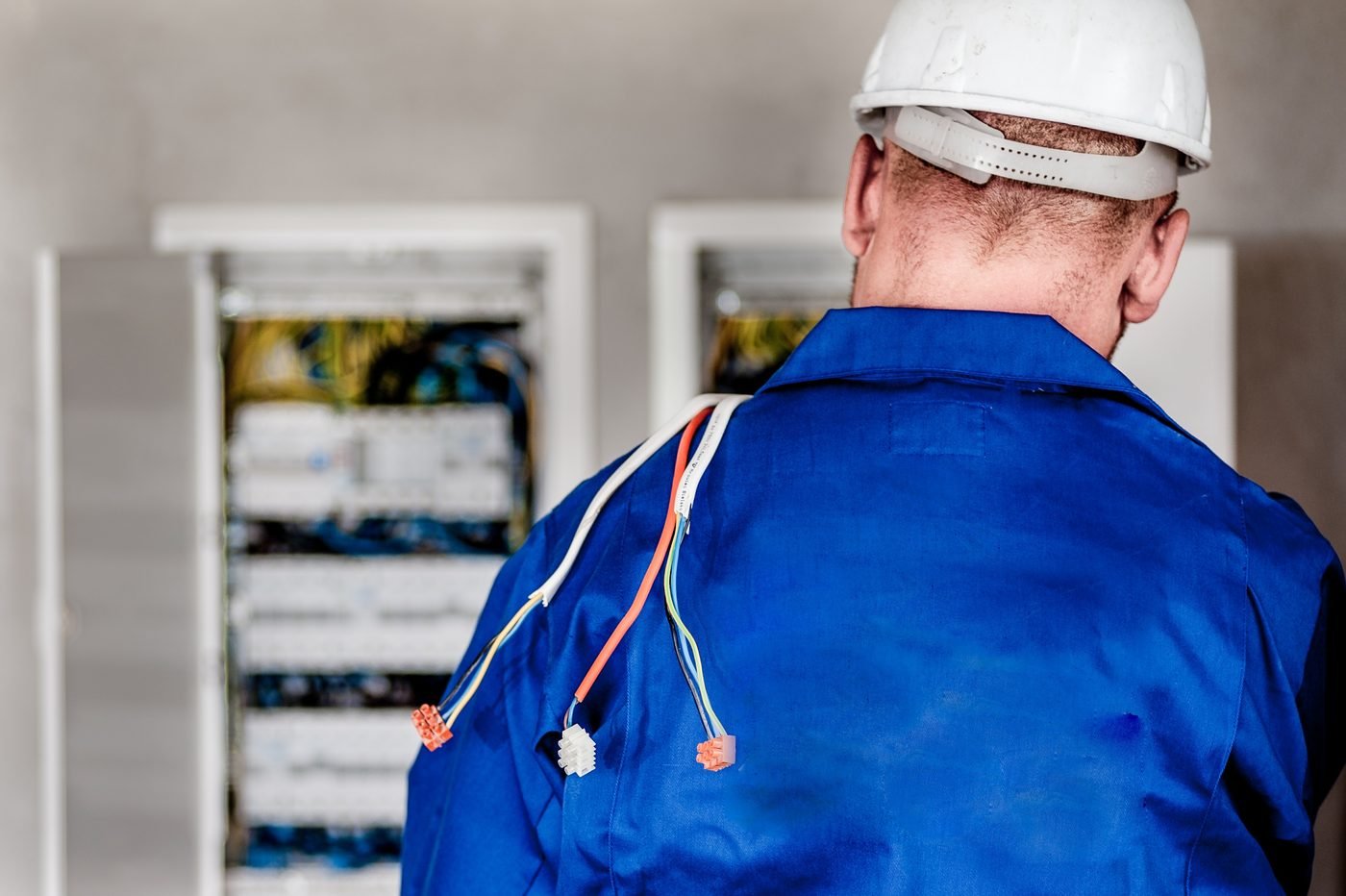 Elektrikas su šalmu ir mėlynu kombinezonu stovi prie atidarytų elektros skirstomųjų spintų