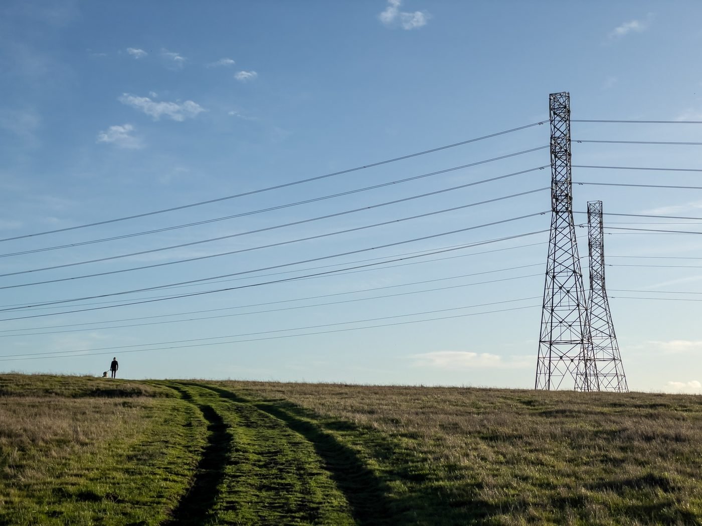 Aukštos įtampos elektros linija virš žolėto lauko, tolumoje einantis žmogus